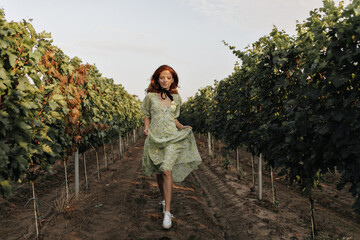 Wall Mural - Full length photo of stylish red-haired girl with black bandage on neck in green summer dress walking on background of vineyards