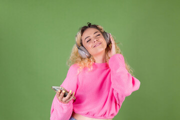 Young blonde woman with long curly hair in pink sweater on green background happy positive enjoying favorite  music in wireless  headphones