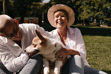 Wall Mural - Blonde lady in hat , white shirt and jeans laughing, sitting on grass with husband in cool eyeglasses and small dog outdoor..