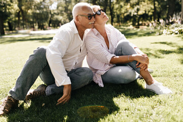 Wall Mural - Handsome man with grey hair in long sleeve shirt and jeans sitting on grass with woman in striped hairstyle and jeans in park..