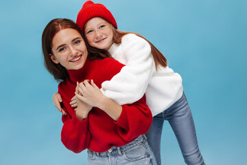 Stylish sisters looking into camera and smiling on blue backdrop. Two girls in modern bright sweaters hugging on isolated background..