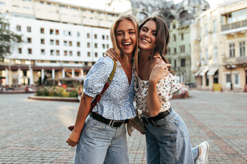 young female friends in stylish jeans and floral trendy blouses hug, smile and pose in great mood at