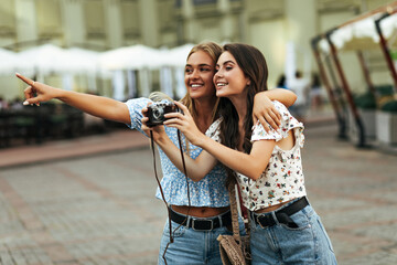 Cheerful brunette and blonde girlfriends in floral cropped blouses and denim pants smiles and walks in city. Attractive girl in blouse top points into distance. Dark haired woman holds retro camera.