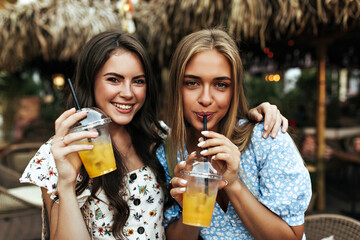  Cheerful brunette curly woman in floral trendy blouse and tanned blonde girl in blue top smile and holds lemonade glasses outside.