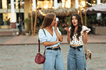 Wall Mural - Curly brunette woman in white floral top and jeans talks to her friend and tells her secrets. Blonde tanned girl in denim pants and blue blouse looks surprised and covers mouth. Women walks in city.