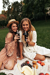 Wall Mural - Pretty brunette curly woman and blonde happy lady in stylish dresses and straw hats smile widely, have picnic in park and drink soda.