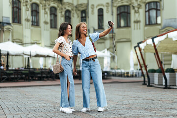 Wall Mural - Attractive young girls in loose stylish denim pants and floral blouses take selfie using retro camera. Blonde and brunette women pose outside.