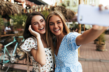 Wall Mural - Cheerful girlfriends in great mood take selfie outdoors and smile. Pretty blonde woman in trendy blue blouse holds cellphone.