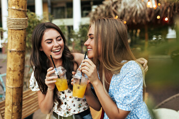 Wall Mural - Good-humored optimistic brunette woman in white floral top and happy blonde tanned girl in blue blouse talk, smile and drink lemonade outside.