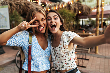 Wall Mural - Tanned blonde girl in blue blouse demonstrates tongue, shows peace sign and winked. Beautiful brunette woman in floral top takes selfie with attractive friend outside.