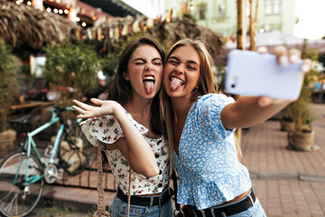 Wall Mural - Active young girls in stylish blouses make funny faces, show tongues and take selfie. Pretty blonde woman in blue top holds purple phone.