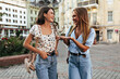 © Look! - Pretty happy brunette and blonde women in stylish denim pants and floral blouses talk. Charming girls in trendy summer outfits walk and smile outside.
