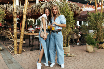 Young optimistic brunette and tanned blonde girls in stylish denim pants and floral trendy summer blouses smile and holds glasses with lemonade.
