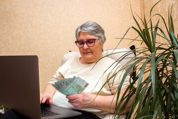 An elderly woman sits in a chair, works with a laptop on her lap and holds money in her hands.