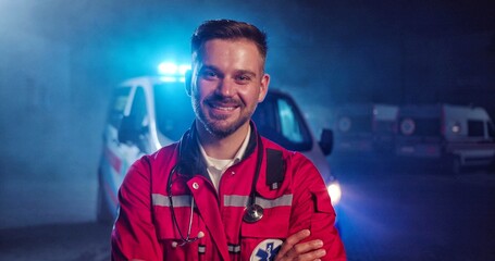 Portrait of Caucasian happy young male paramedic in red uniform smiling to camera and standing outdoor. Ambulance car on background. Attractive cherful male doctor at night shift. Call 911