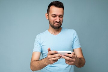 Photo of asking handsome young man with beard wearing everyday blue t-shirt isolated over blue background holding and using mobile phone communication online on the internet looking at gadjet display
