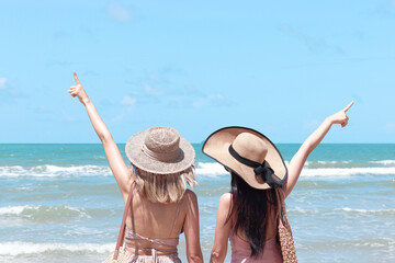 Two beautiful Asian women with big hat from behind enjoy spending time on tropical sand beach blue sea together, resting and relaxing on summer holiday vacation.