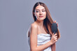 © Olena Apetroaiei - A beautiful brown-haired woman with bared shoulders looks at the camera as she brushes her hair with a brush, isolated on a gray background with copy space for text.