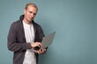 © Ivan Traimak - Photo of handsome blond man holding computer laptop looking at camera isolated over blue wall background wearing white t-shirt and grey sweater