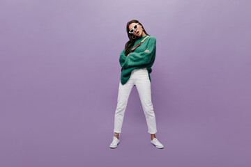 Wall Mural - Serious brunette woman in white pants and oversized green sweater poses on purple background. Full-length portrait of charming girl in sunglasses on isolated.