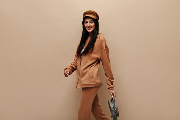 Wall Mural - Tanned brunette woman in light brown suit and cap posing on isolated. Charming girl holds grey handbag on beige background.