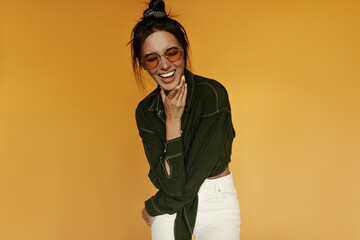 Wall Mural - Charming woman in white pants and dark green shirt laughs on isolated. Brunette girl in orange sunglasses smiles and poses on yellow background.