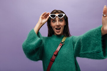 Wall Mural - Surprised green-eyed tanned woman takes off sunglasses. Cute shocked lady in stylish woolen sweater makes selfie on purple background.