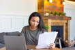 © Stavros - Asian woman works with documents in front of a laptop monitor. She checks the tax records before submitting the report.