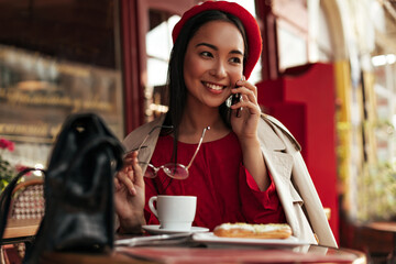 Wall Mural - Portrait of tanned brunette Asian woman in beige trench coat, red beret and dress holds eyeglasses, talks on phone and enjoys coffee in street cafe.