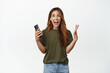 Cheerful middle aged woman holding mobile phone, looking at camera and screaming with rejoice, winning online smartphone, standing against white background