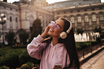 Wall Mural - Charming brunette woman in stylish hoodie and pink sunglasses enjoys music in headphones. Young girl smiles sincerely outside.