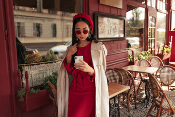 Wall Mural - Elegant charming brunette Asian woman in stylish red dress, beret, sunglasses and trench coat holds cellphone and leans on wooden table in street cafe.