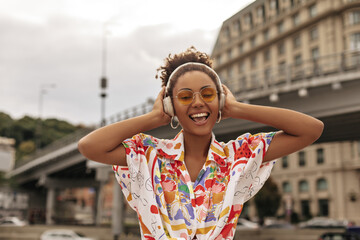 Beautiful young curly brunette dark-skinned woman in colorful blouse and orange sunglasses listens to music in headphones and sings outside.