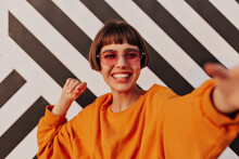 Cheerful Short-haired Girl In Bright Clothes Smiling Outside. Modern Woman In Sunglasses Making Selfie On Striped Backdrop..
