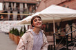 Short-haired girl in pink jacket walks outdoors. Cheerful lady in light outfit smiling and looking into camera at street..