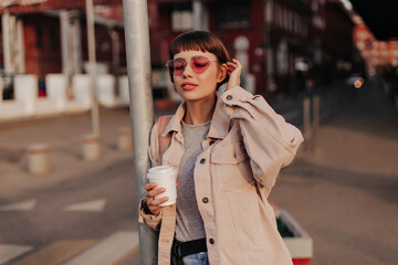 Wall Mural - Fashionable lady holding cup of coffee outside. Brunette girl in denim beige jacket and pink glasses posing with closed eyes in city..