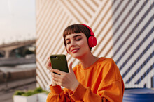 Charming Lady With Brunette Hair In Red Headphones Holding Phone Outside. Girl In Orange Sweatshirt Listening To Music On Striped Backdrop..