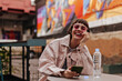 © Look! - Cheerful woman with brunette hair laughing outside. Modern short-haired girl in bright glasses and denim light outfit holding phone in cafe..