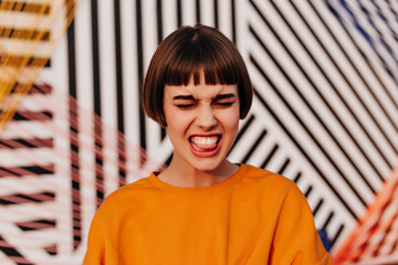 Wall Mural - Emotional girl with brunette hair in orange sweatshirt makes funny face outside. Hipster in bright clothes showing tongue on striped backdrop..