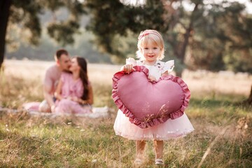 Wall Mural - Girl are dressed in pink dress and holding big pink heart in hands. Mom, dad are sitting next to wigwam decoration in the park. Family spending time outdoor in summer, having fun together.