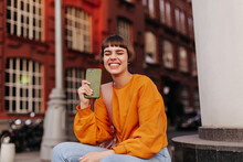 Joyful Short-haired Woman In Orange Sweatshirt Smiles With Closed Eyes Outside. Happy Girl Holds Smartphone In Khaki Case.