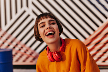 Wall Mural - Optimistic brunette woman in orange clothes laughing in cafe. Short-haired girl with red headphones posing on striped background..