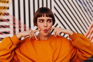 Wall Mural - Funny girl with short hairstyle in bright outfit has fun outdoors. Modern woman in orange modern sweatshirt posing on striped backdrop..