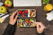 First person top view photo of hands eating healthy food salad berries nuts from two lunchboxes apples glass with juice plant stationery keyboard and mouse on isolated dark wooden desk background