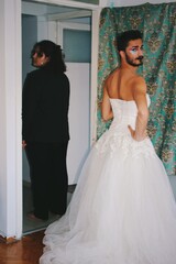 portrait of queer young man with beard & body hair in white bridal gown & young woman in black suit / wedding cross dressing at home