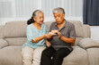© ฺฺฺBoonterm - Elderly Asian couple sitting on the sofa eating donuts in the living room together : Eating sweets for the elderly and taking care of the health of the elderly in retirement concept
