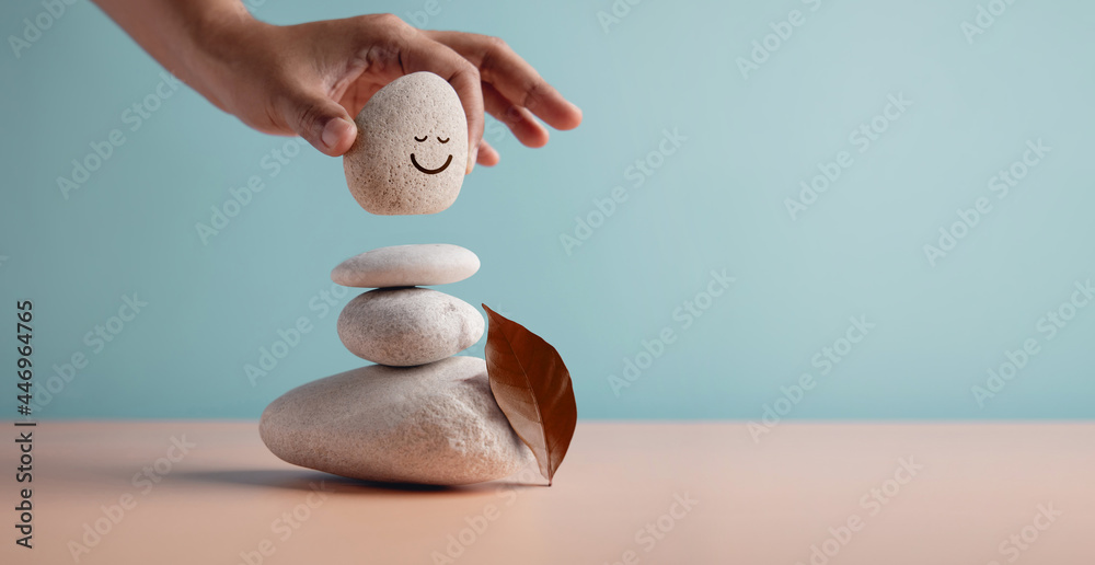 Enjoying Life Concept. Harmony and Positive Mind. Hand Setting Natural Pebble Stone with Smiling Face Cartoon to Balance on Beach Sand. Balancing Body, Mind, Soul and Spirit. Mental Health Practice - obrazy, fototapety, plakaty 