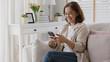 © ChayTee - Old adult asia female sit relax on sofa couch smile watch, play, text chat on cellphone at home in happy retired in older people, elderly health care tech life, enjoy pay money online via banking app.