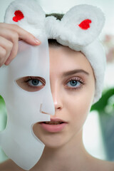 Wall Mural - Young woman holds facial mask on half of her face. Spa and Wellness, Skin Care Concept. Close-up, selective focus