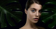Young woman with perfect skin arounded by tropical green leaves on dark background. Beautiful fashion model with monstera plants. Concept of natural organic cosmetics, 